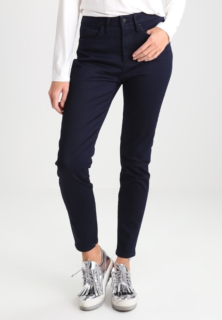 Personne portant un pantalon slim bleu marine foncé et des chaussures à plateforme argentées brillantes à lacets, debout avec une main reposant sur la hanche.