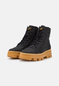 Bottes en tissu noir avec une semelle en caoutchouc, présentant un bout rond, un laçage à l'avant et une semelle extérieure de couleur gomme contrastante.