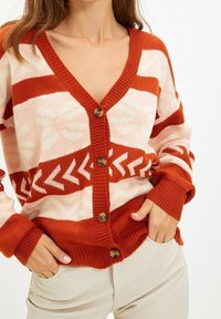 Gestrickte Strickjacke in Rost und Creme mit V-Ausschnitt, geometrischen Mustern, fünf braunen Knöpfen sowie gerippten Bündchen und Saum.