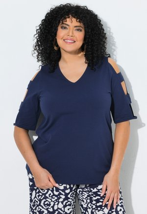 Frau mit lockigem schwarzen Haar lächelt, trägt ein navyblaues Cold-Shoulder-Oberteil und gemusterte schwarz-weiße Hose vor weißem Hintergrund.
