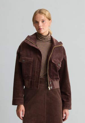 Frau mit blond gebundenem Haar, die eine braune Cordjacke über einem karierten Rollkragenpullover und einem passenden braunen Cordrock trägt.