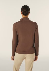 Top marrone a maniche lunghe con una texture morbida e un colletto, caratterizzato da un design aderente che si stringe in vita, abbinato a pantaloni beige chiaro.
