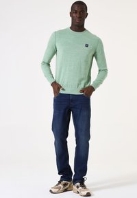 Lichtgroene trui met een ronde hals, gecombineerd met donkerblauwe jeans en beige sneakers. De trui heeft een klein logo op de borst.
