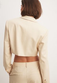 Beige cropped blazer met een gladde textuur, met schouderflappen, lange mouwen en knoopmanschetten. Gecombineerd met bijpassende geklede broek.