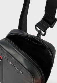 Bolso de hombro negro con cremallera, hecho de material liso, con un logo en la parte frontal, correa ajustable y forro interior suave.