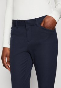 Närbild av person som bär en vit långärmad tröja och marinblå byxor med handen i fickan, som visar tygtextur och passform.