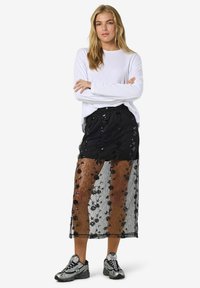 Gonna maxi nera in pizzo con motivi floreali e paillettes, sovrapposta a un tessuto nero solido. Abbinata a una maglietta bianca a maniche lunghe e sneakers.