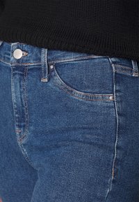 Mellanlånga blå denimjeans med en klassisk femfickdesign, med silverdetaljer och kontrasterande sömmar. Texturerad tygsynligt.