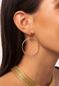 Goldene Creolen mit einem kleinen roten Steinakzent, die ein glattes, abgerundetes Design aufweisen. Die Textur ist glänzend und reflektierend, was das Erscheinungsbild verstärkt.
