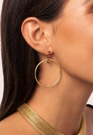 Goldene Creolen mit einem kleinen roten Steinakzent, die ein glattes, abgerundetes Design aufweisen. Die Textur ist glänzend und reflektierend, was das Erscheinungsbild verstärkt.