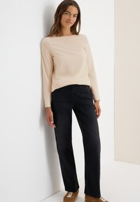 Light beige long-sleeve top with a textured pattern paired with dark, relaxed-fit jeans. Model stands against a white background.