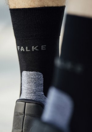 Schwarze und graue Falke-Socke, getragen mit einem schwarzen Schuh, mit dem Markennamen der Socke auf der Rückseite des Knöchels sichtbar.
