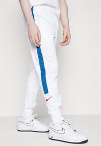Pantalons de sport blancs avec une bande latérale bleue, dotés d'une taille élastique et d'un logo Nike rouge près de la cheville. Associés à des baskets blanches.