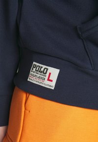 Marinblå hoodie med ribbade ärmslut, tillsammans med orangea byxor. Lappen läser "POLO SPORTING GOODS RALPH LAUREN KVALITET ATLETISK KLÄDSEL."