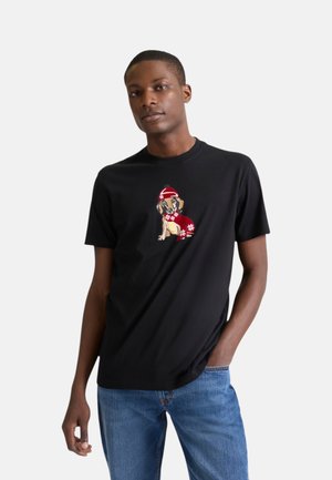 Giovane uomo che indossa una t-shirt nera con una piccola grafica di un cane con cappello e sciarpa rossi invernali, abbinata a jeans blu.