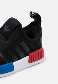 Svart sportskor med övre del av textil, med tre svarta ränder, röda och blå detaljer på en vit sula, och en dragflik vid hälen.