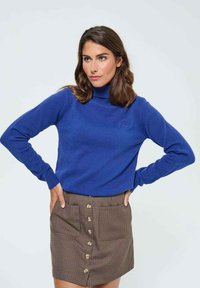 Pull en col roulé bleu en matière douce, à manches longues. Assortie à une mini-jupe à carreaux marron avec des détails de boutons sur le devant.