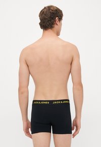 Mann von hinten gesehen, der schwarze Boxershorts mit schwarzem Bund trägt, auf dem gelber Text "JACK & JONES" steht.
