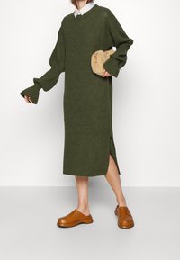 Vihreä neulemekko, jossa on V-pääntie, pitkät liehuvat hihat, sivuvaste ja beige pörröinen laukku. Mallilla on jalassaan ruskeat puukengät.