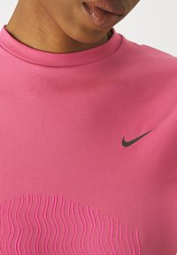 T-shirt de sport Nike rose avec un col montant et une texture à motif ondulé subtil. Présente un logo noir sur la poitrine gauche.