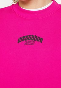Gros plan d'une personne portant une chemise rose vif avec le texte noir « westblue studio » imprimé sur la poitrine.