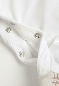 Tissu en coton blanc avec des pressions métalliques sur un côté et un détail brodé beige clair. Le matériau est doux avec un léger éclat.