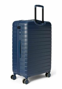 Valise rigide bleu foncé avec texture côtelée verticale, poignée noire rétractable et quatre roues pivotantes pour un transport facile.