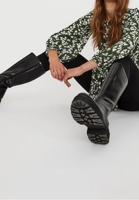 Bottes noires montant jusqu'aux genoux avec une finition lisse et des semelles texturées, associées à un chemisier vert à motifs floraux et un pantalon noir, posées sur un fond neutre.