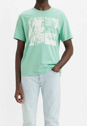 Bavlněné tričko světle zelené barvy s abstraktními bílými grafickými potisky. Krátké rukávy, kulatý výstřih, volný střih, nošeno s světle modrými džínami.