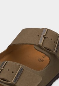 Καφέ δερμάτινο σανδάλι με δύο ρυθμιζόμενες μεταλλικές αγκράφες με την επιγραφή "Shepherd" και μαλακό ταμπά πάτο.