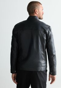 Chaqueta de cuero negra con un diseño ajustado, que presenta un cuello alzado, acentos cosidos y puños abotonados, vista desde la parte trasera.