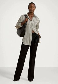 Sieviete, tērpusies brīvā svītraina pogu krekla, melnās plašās biksēs un nēsājot lielu melnu plecu somu, stāv uz vienveida fona.