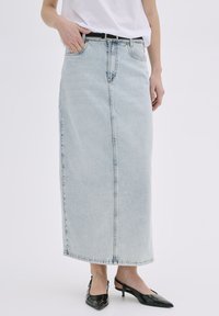 Moteris dėvi šviesiai mėlyną aukštai liemeniai džinsų maxi sijoną, baltą marškinėlį, juodą diržą ir juodus smailius slingback batus, stovinti baltoje fone.