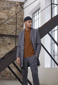 Manteau en mélange de laine gris sur un pull en tricot marron, associé à un pantalon à carreaux gris. Textures douces et coupes nettes sont mises en avant.