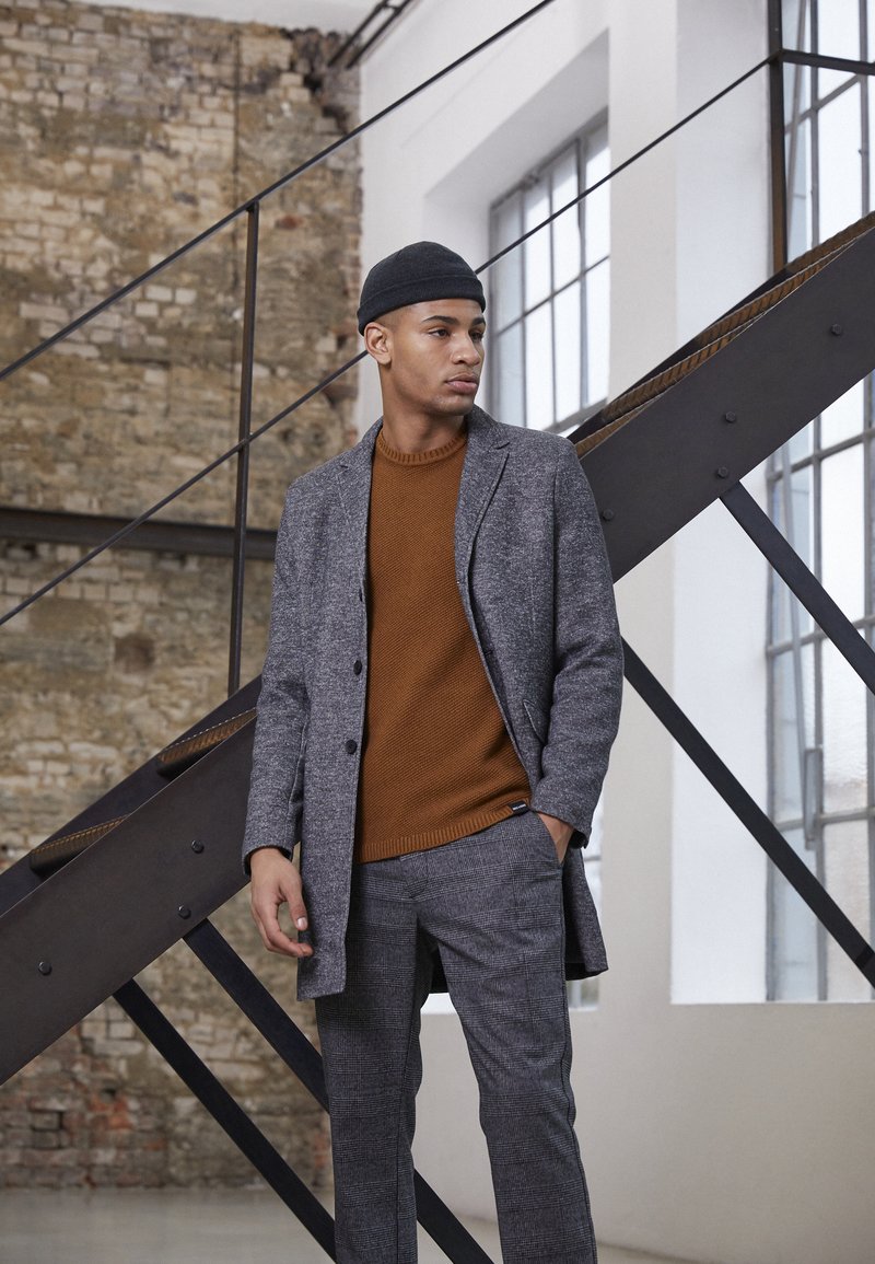 Manteau en mélange de laine gris sur un pull en tricot marron, associé à un pantalon à carreaux gris. Textures douces et coupes nettes sont mises en avant.