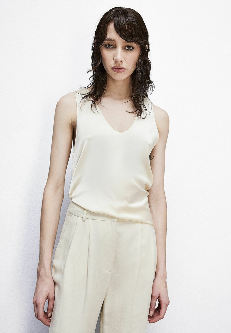 Top in satin con scollo a V di un morbido crema, abbinato a pantaloni beige chiaro su misura. Texture liscia, design minimale e senza motivi visibili.