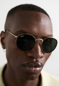 Ochelari de soare rotunzi, negri, cu ramă din metal aurit și detalii pe punte. Logo-ul „Ray-Ban” pe lentilă, finisaj lucios, purtați de o persoană.