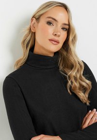 Pull à col roulé noir avec une finition texturée, manches longues et coupe ajustée. Le modèle porte des cheveux ondulés de longueur épaule.