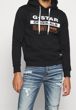 Osoba nosząca czarną bluzę z kapturem G-Star Originals Raw z denimu i podarte niebieskie dżinsy, stoi na jednolitym jasnym tle.