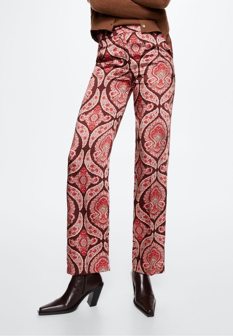 Pantalons marron à motifs présentant un design paisley en rouge et crème. Jambe droite ajustée avec une taille mi-haute. Associés à des bottes à talons sombres.