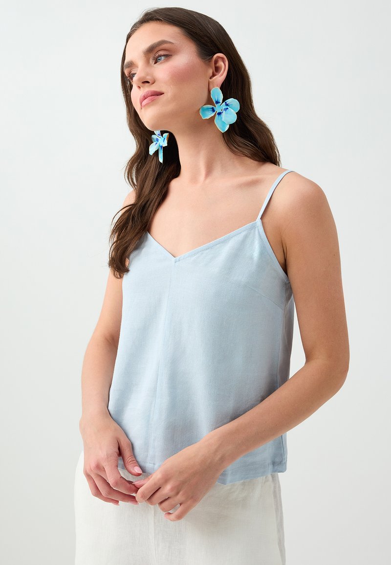 Femme aux longs cheveux bruns portant un top bleu clair style camisole et de grandes boucles d’oreilles bleues en forme de fleur sur un fond uni.