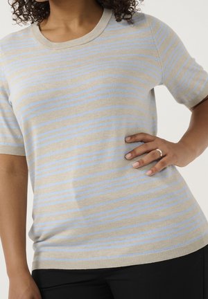 Frau trägt beige und hellblau gestreiftes kurzärmliges Stricktop, schwarze Hose, mit der Hand auf der Hüfte, die einen silbernen Ring zeigt.