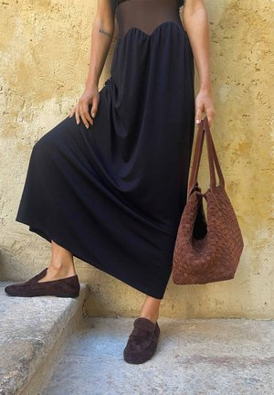 Personne portant une longue robe noire et des mocassins marron, tenant un grand sac à main tressé marron, debout sur des marches en pierre près d'un mur texturé.