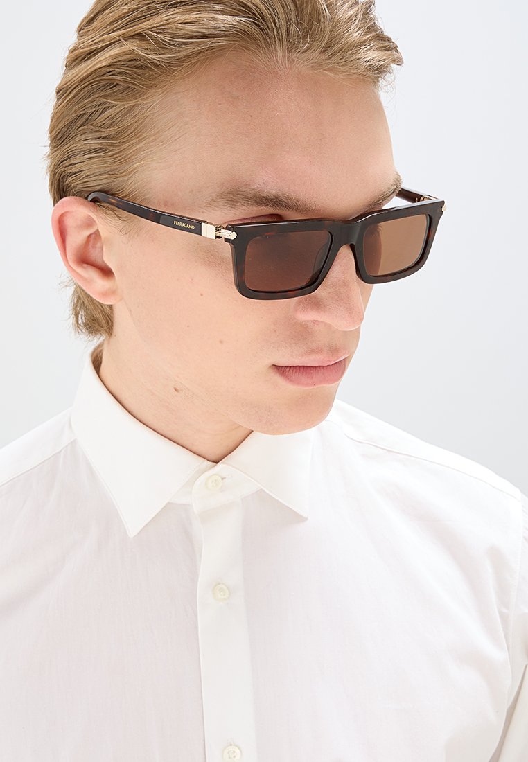 Lunettes de soleil noires rectangulaires avec des verres teintés bruns, dotées d'un accent doré sur la branche et d'un logo subtil, portées avec une chemise blanche à boutons.
