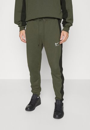 Mann trägt olivgrüne Nike Air Jogginghose mit schwarzen Seitenpaneelen, passendem Sweatshirt und schwarzen High-Top-Sneakers vor neutralem Hintergrund.