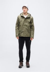 Αδιάβροχο μπουφάν χακί ελιάς με κουκούλα, φερμουάρ μπροστά και πολλαπλές τσέπες, συνδυασμένο με μαύρο παντελόνι και καφέ παπούτσια πεζοπορίας.