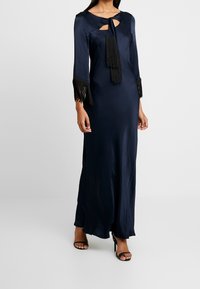 Vestido maxi de satén navy con mangas largas, escote con ojo de llave, acentos de flecos negros en las mangas y textura suave y fluida.