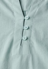 Camicia azzurra chiara con scollo a v, quattro bottoni circolari e una piccola tasca sul petto. Il tessuto ha una finitura liscia e leggermente strutturata.