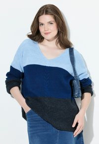 Junge Frau mit schulterlangem braunem Haar, die einen farblich blockierten blauen Pullover und einen Jeansrock trägt, während sie eine kleine blaue Schultertasche trägt.