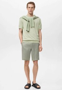 Pulover scurt cu mâneci scurte de culoare verde deschis, cu o textură ribbed, asortat cu pantaloni scurți de aceeași culoare. Sandalele negre completează ținuta, prezentată pe un fundal neutru.